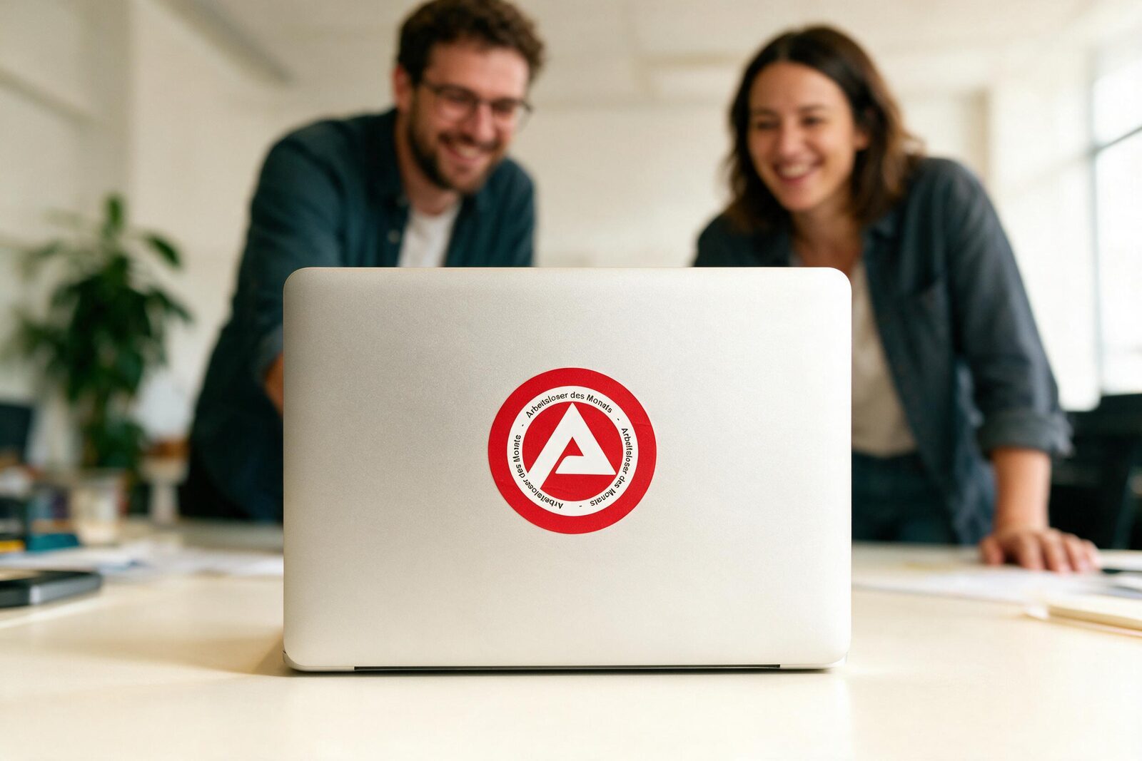Laptop mit dem runden Arbeitsagentur-Parodie-Sticker, dahinter grinsende Kollegen